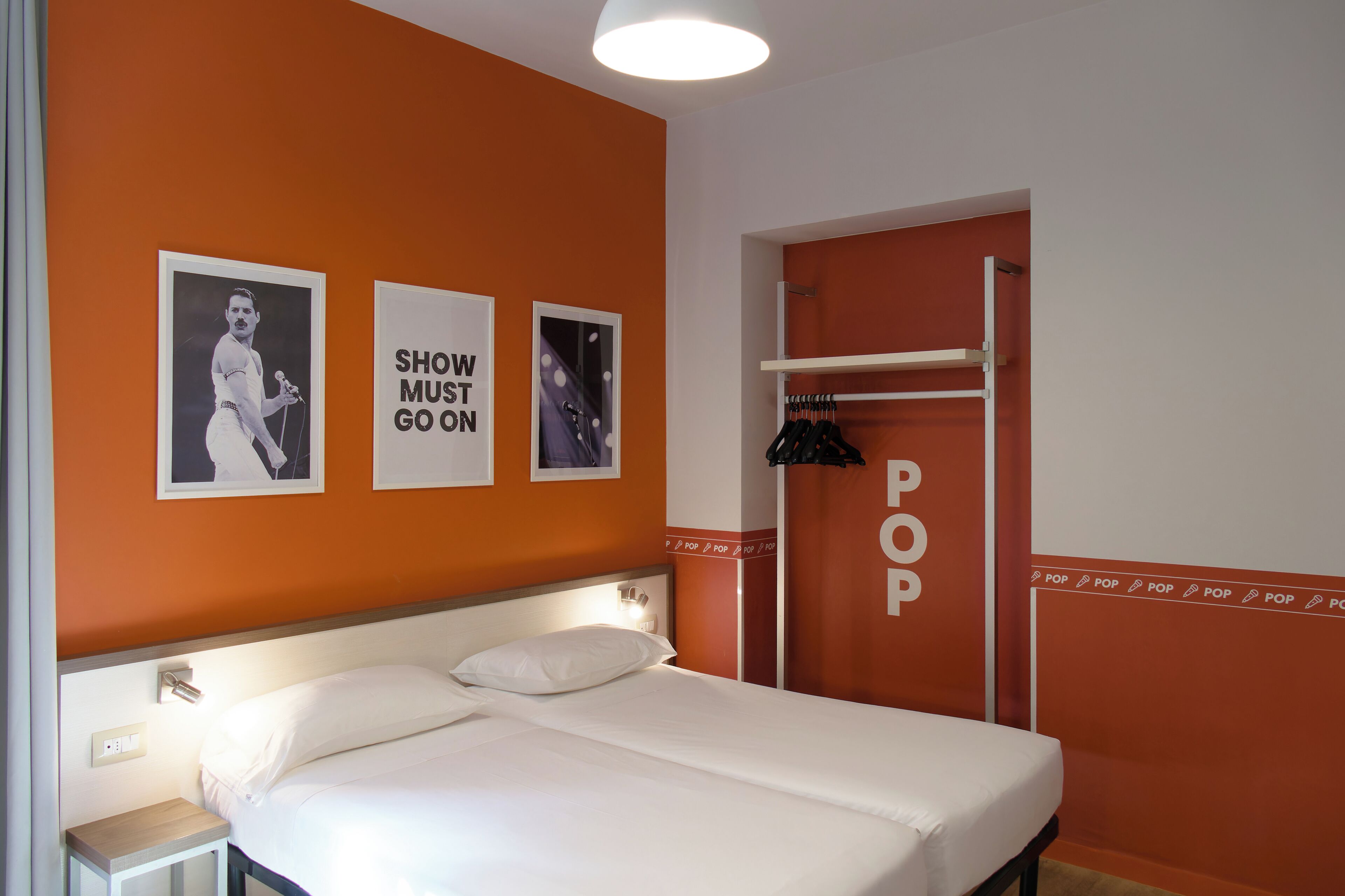 Une chambre d’hôtel avec un lit, une table de chevet et une armoire. Il y a des affiches au mur et une pancarte sur laquelle on peut lire « POP ».