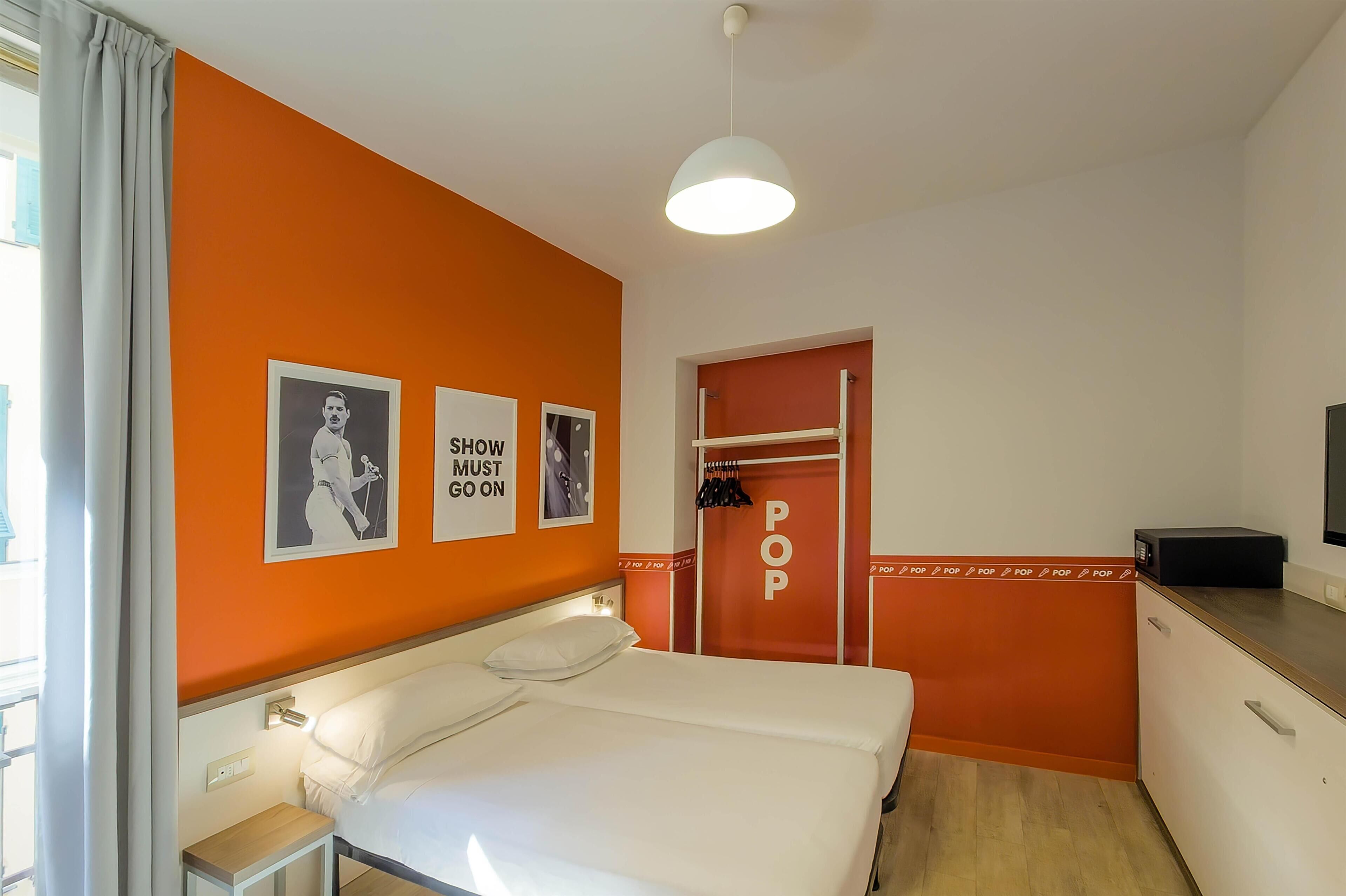 Habitación de hotel con pared de acento naranja, cama con ropa de cama blanca, mesita de noche pequeña, puerta roja con letrero 'POP' y lámpara colgante blanca.