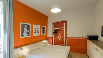Ein Hotelzimmer mit orangefarbener Akzentwand, einem Bett mit weißer Bettwäsche, einem kleinen Nachttisch, einer roten Tür mit der Aufschrift 'POP' und einer weißen Pendelleuchte.