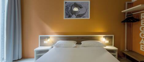 Habitación de hotel con una cama grande, dos mesitas de noche, una obra de arte en la pared y un perchero.