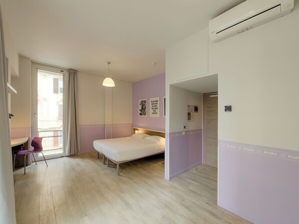 Habitación de hotel con cama, escritorio, silla, ventana con cortinas, aire acondicionado de pared y puerta que da a otra habitación.