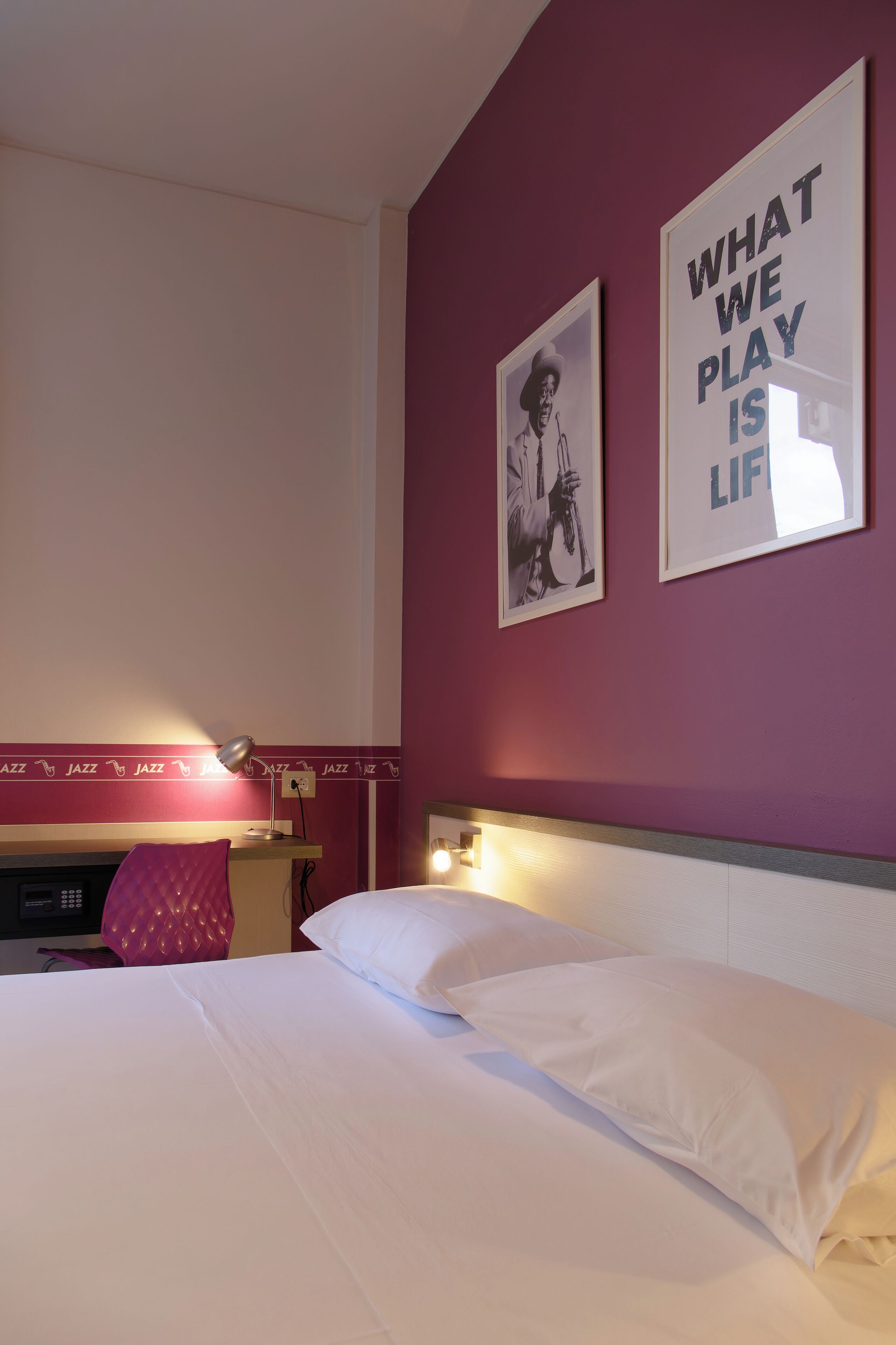Ein Hotelzimmer mit einem Bett, einem Schreibtisch und einem Stuhl. An der Wand hängen zwei gerahmte Bilder.