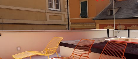 Una terrazza con sedie e tavolo in metallo arancione, riparata da un ombrellone dello stesso colore, con vista su un edificio con tetto in tegole e una finestra con persiane verdi.
