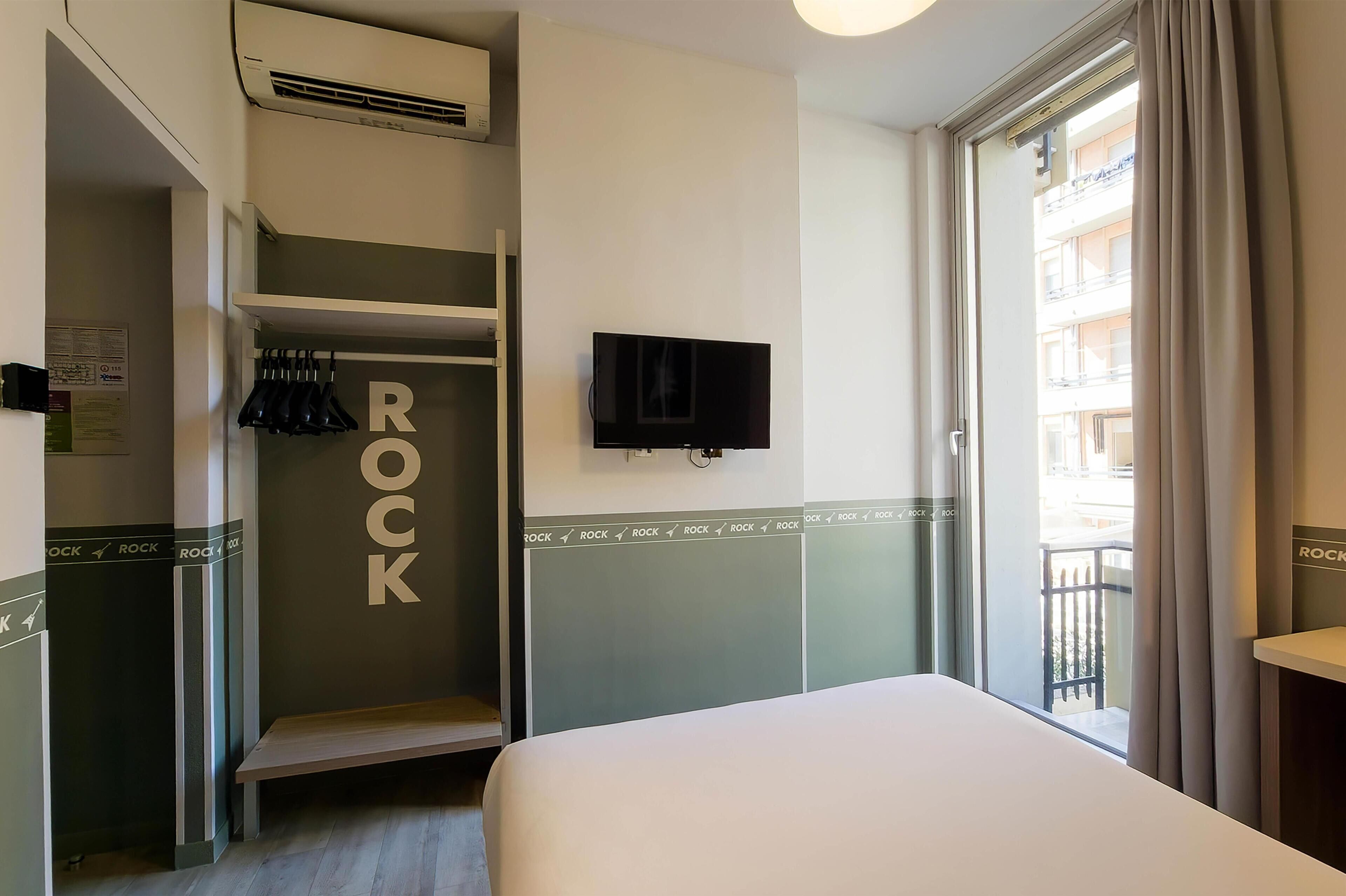 Ein Hotelzimmer mit Bett, Wandfernseher, Kleiderschrank und Balkon mit Blick auf Gebäude.