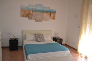 Un dormitorio con una cama, dos mesitas de noche, una mesilla auxiliar, una obra de arte en la pared y una ventana con cortinas.