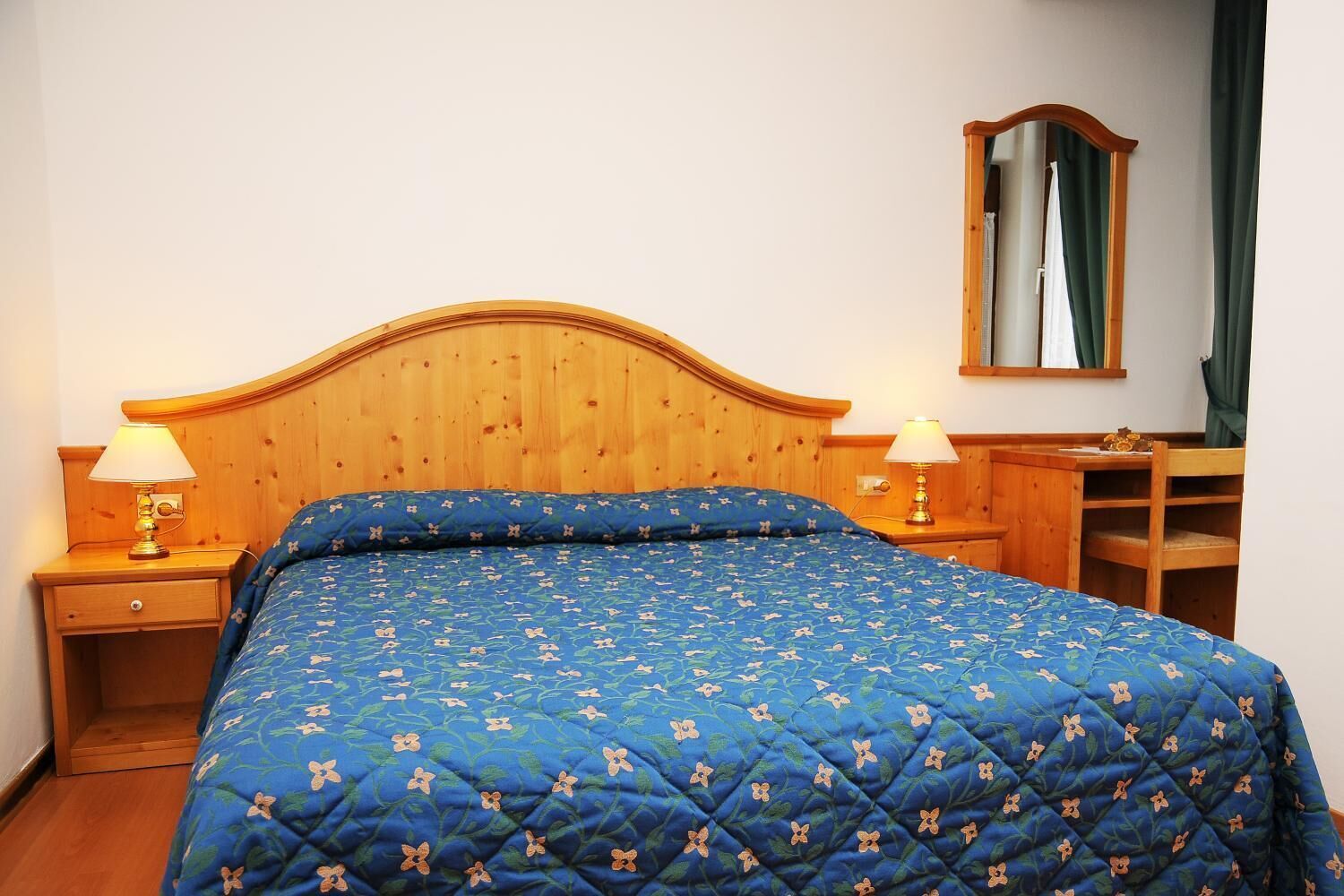 A bedroom with a wooden headboard, a blue floral bedspread, two bedside lamps, a wooden nightstand, a mirror, and a wooden dresser.