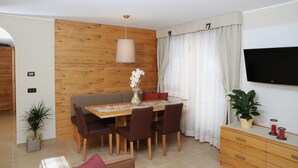 A dining area with a wooden wall, a table with chairs, a vase with flowers, a television on the wall, and a plant on a dresser. - Residence Marisol (Mezzana)