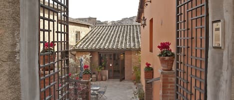 A courtyard with a tiled roof, a brick wall, and a metal gate. There are potted plants and a table with chairs.