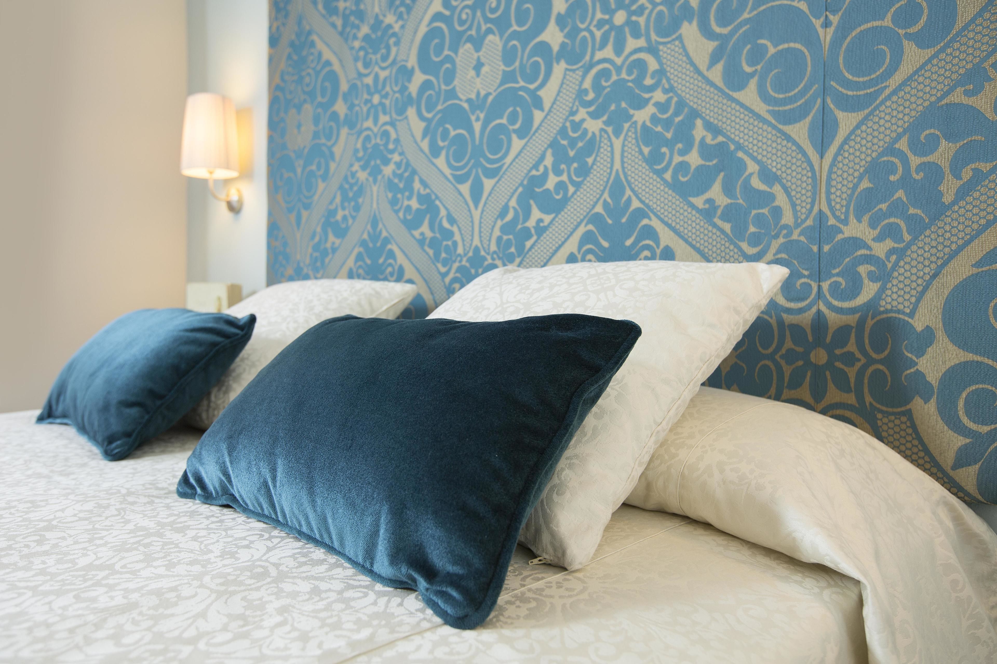 A bed with patterned bedding and pillows, a wall with a blue and beige design, and a wall-mounted light fixture.