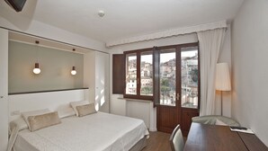Un dormitorio con cama, mesita de noche, lámpara y ventana con vistas a edificios.