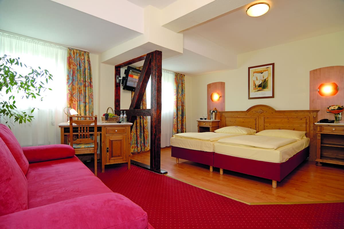 Une chambre d’hôtel avec un lit, un bureau, une chaise, un canapé rouge et une fenêtre avec des rideaux.
