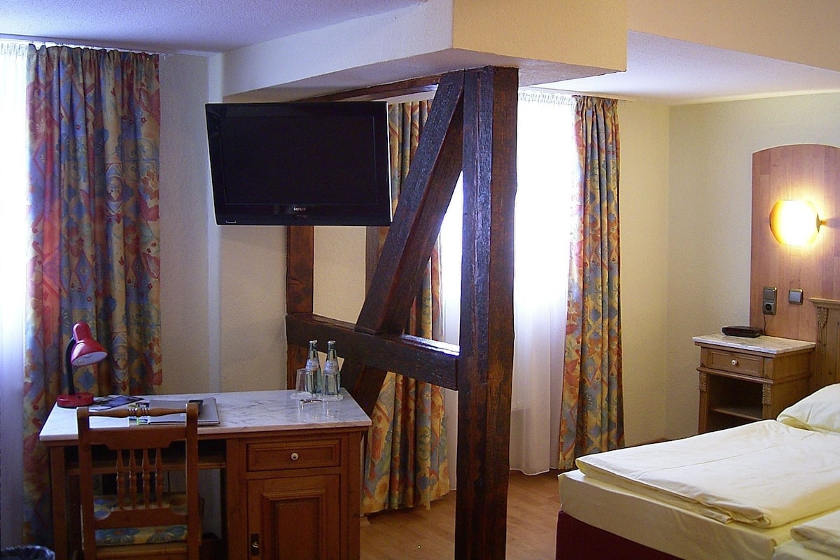 Une chambre d’hôtel avec un lit à baldaquin en bois, un bureau avec une chaise, un téléviseur fixé au mur et une table de chevet avec une lampe.