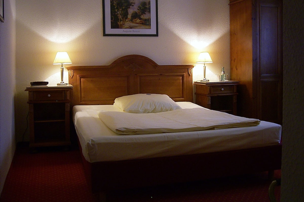 Un lit bien fait, avec deux tables de chevet, chacune surmontée d’une lampe, dans une chambre d’hôtel.