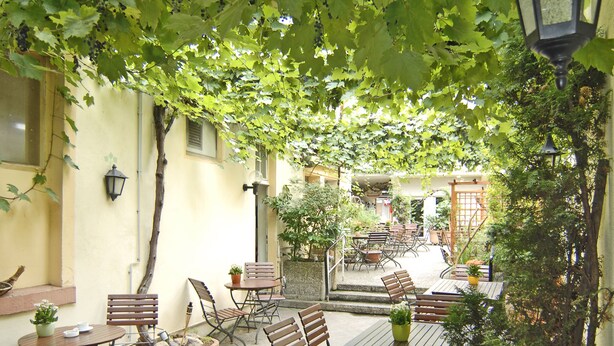 An outdoor patio area with wooden tables and chairs, surrounded by potted plants and a hanging lantern. - Hotel Restaurant Bockshaut (Darmstadt)