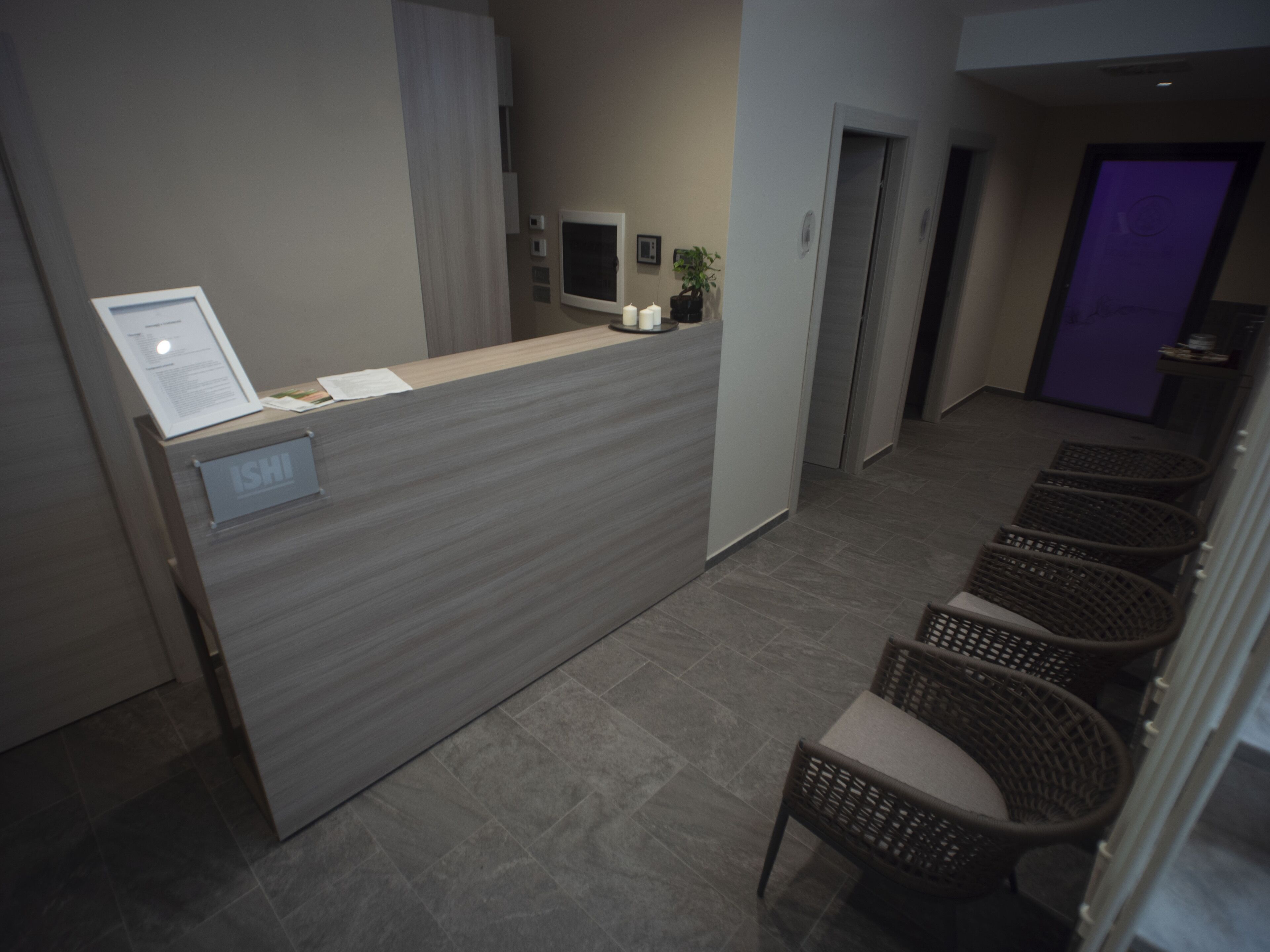 A reception desk with a sign reading "ISHI" and a computer monitor. There are wicker chairs and a door in the background.