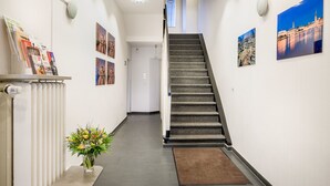 Een gang met een tapijtvloer, een wenteltrap, een radiator, een bloemenvaas en lijstjes met afbeeldingen aan de muur.