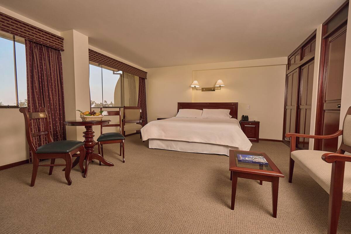 Une chambre d’hôtel avec un grand lit, deux tables de chevet, une chaise, une petite table avec un panier, et une vue sur l’extérieur.