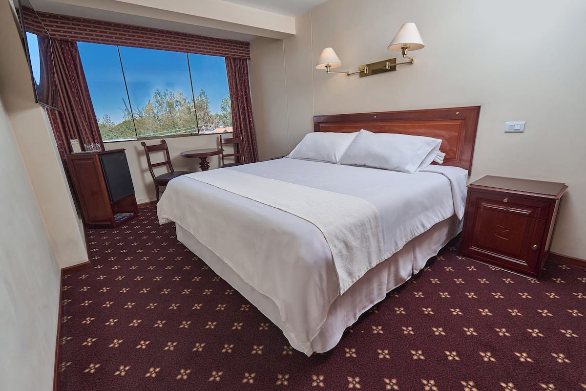 Une chambre d’hôtel avec un grand lit, un bureau, une chaise, une petite table, une table de chevet et une fenêtre avec des rideaux.
