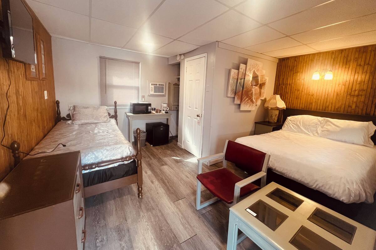 Une chambre d’hôtel compacte comprenant deux lits simples, un bureau avec un ordinateur et un micro-ondes, un petit coin salon et un mur décoratif en bois.