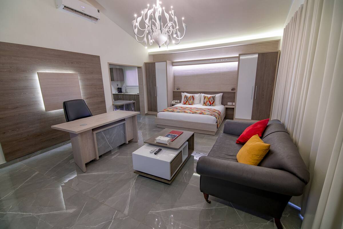 A modern hotel room with a desk, a bed with colorful pillows, a gray sofa with red and yellow cushions, and a chandelier.