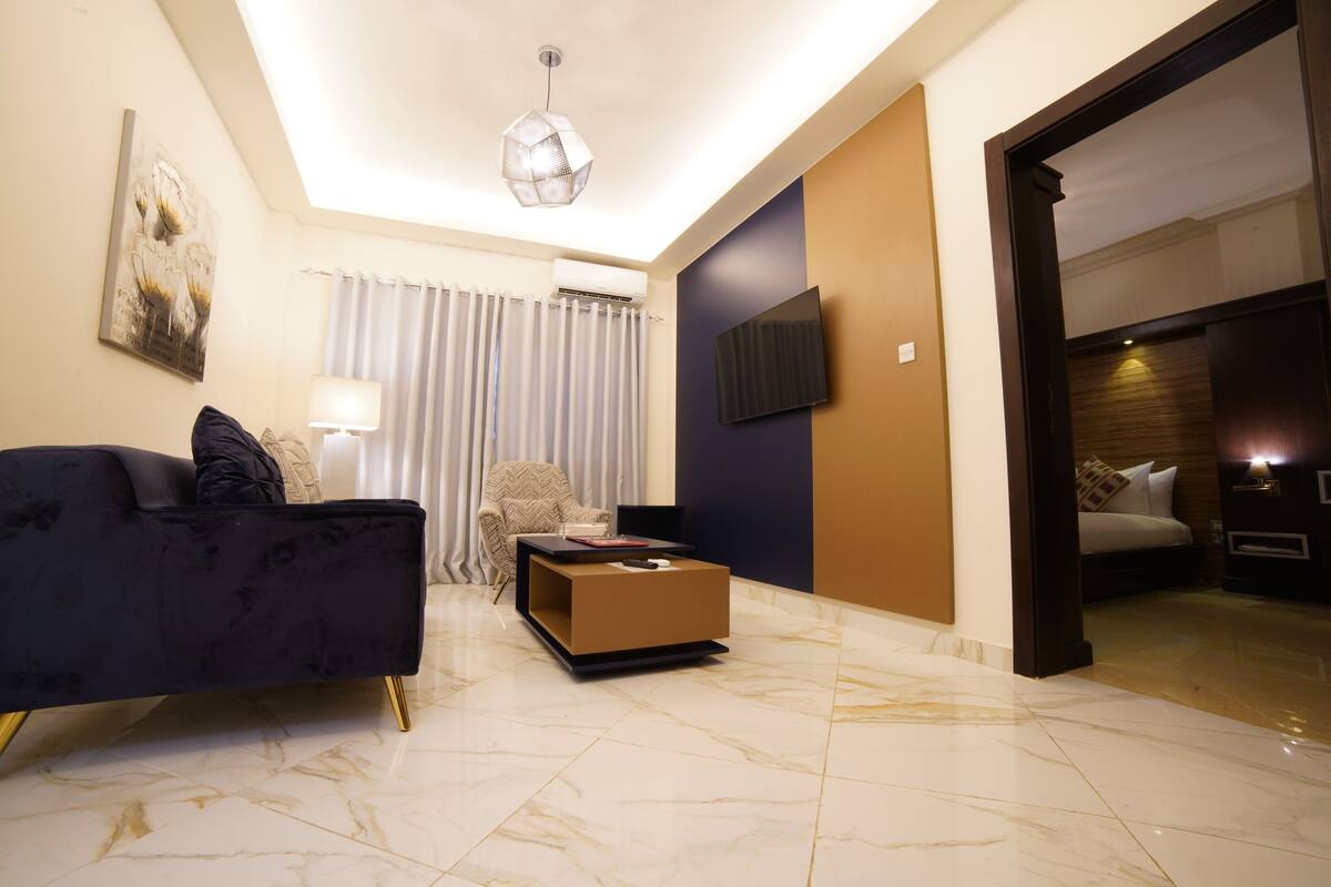 A modern hotel room with a dark blue sofa, a small wooden coffee table, a wall-mounted TV, and a large window with curtains.