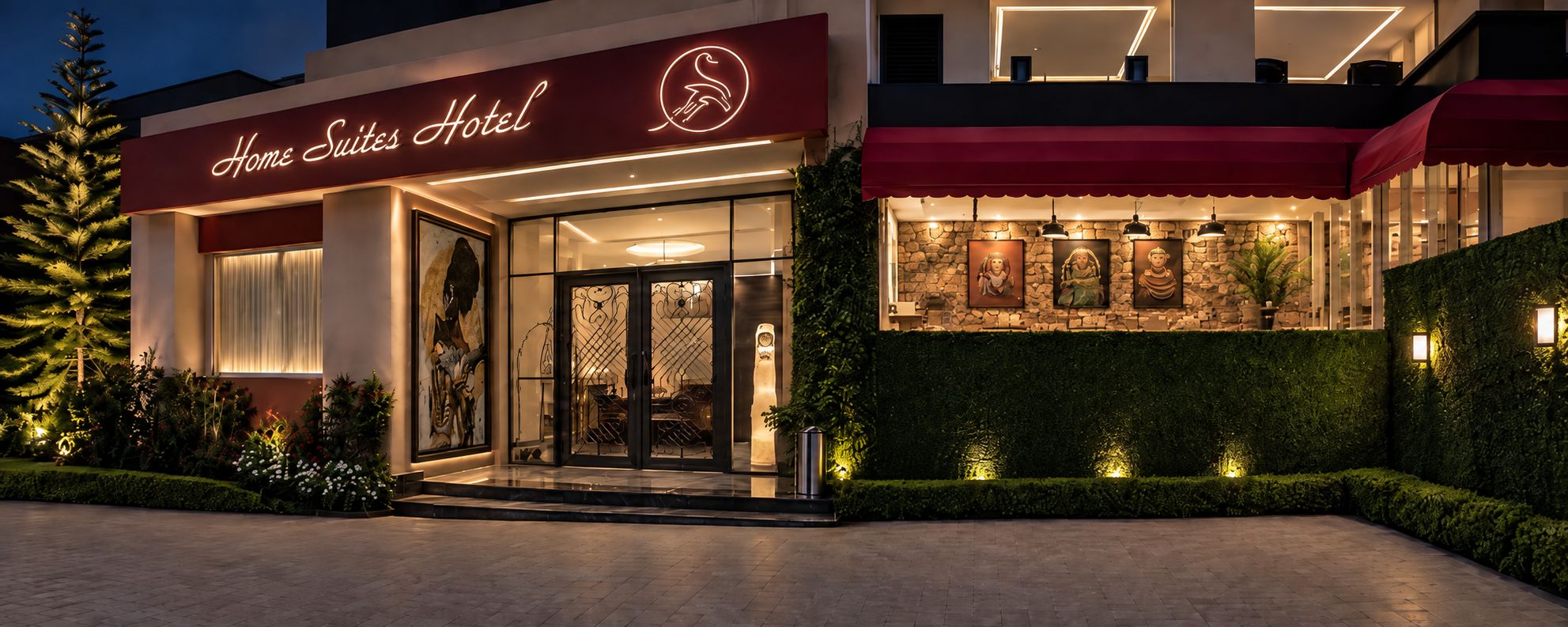 The exterior of a hotel with a neon sign, a glass entrance, and a landscaped garden.