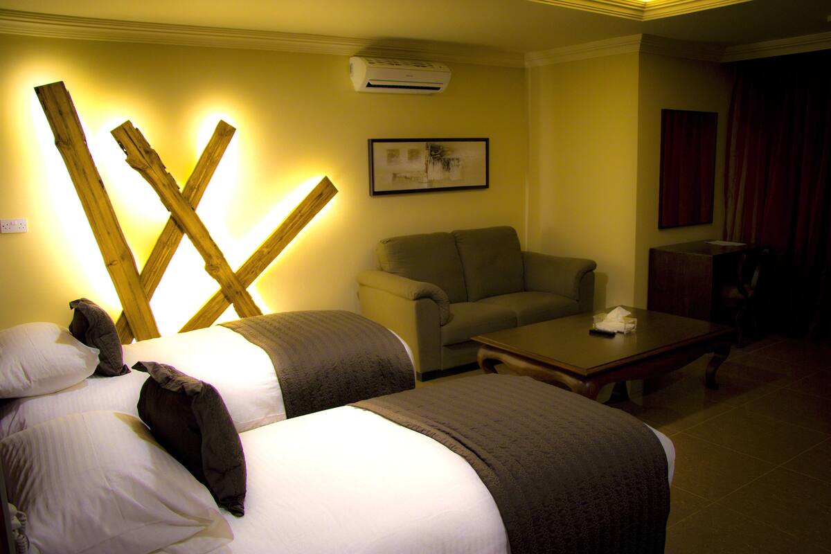 A hotel room with two beds, a sofa, a table, and an air conditioning unit.
