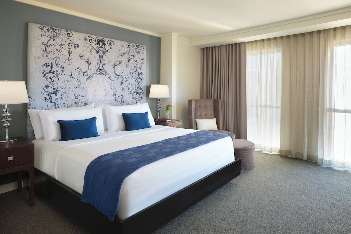 A large bed with a blue blanket, two bedside tables with lamps, a chair, and a large window with curtains.