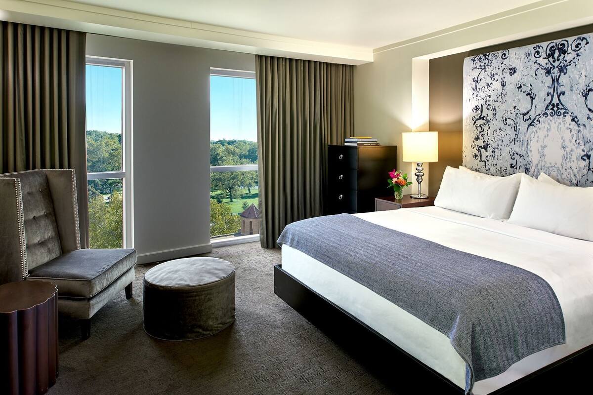 A hotel room with a large bed, a chair, a small ottoman, a nightstand, a lamp, and a view of greenery outside.