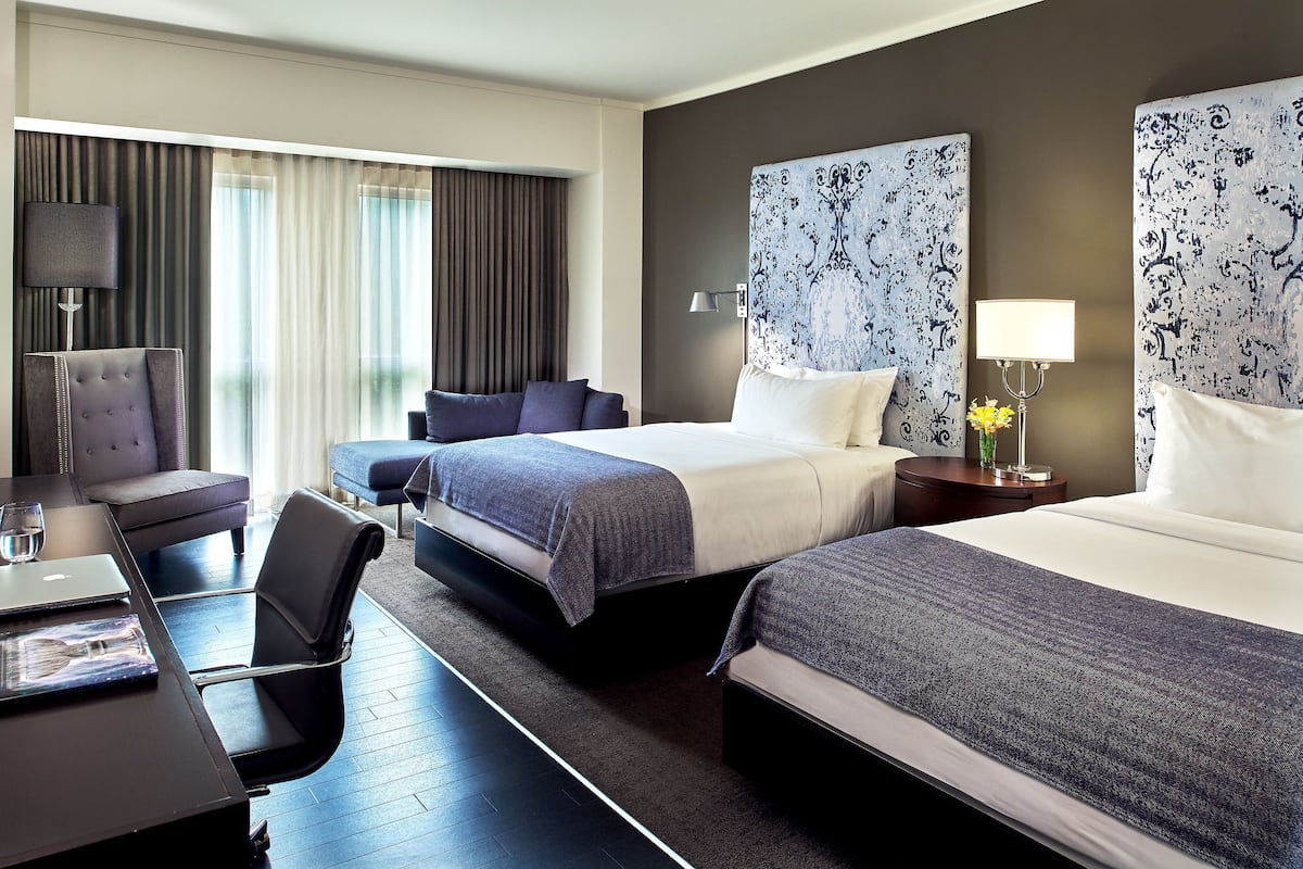A hotel room with two beds, a desk with a laptop, a chair, and a large window with curtains.