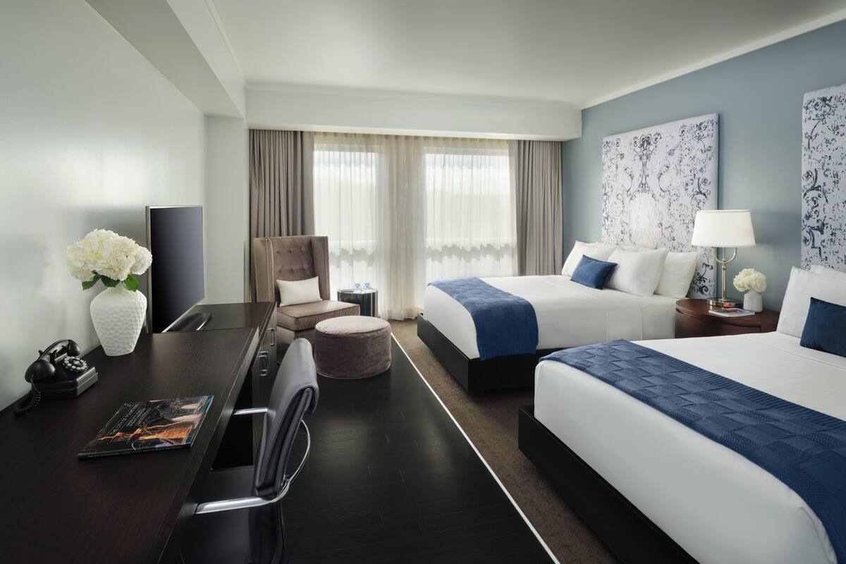 A hotel room with a desk, two beds, a chair, a round ottoman, a lamp, and artwork on the walls.