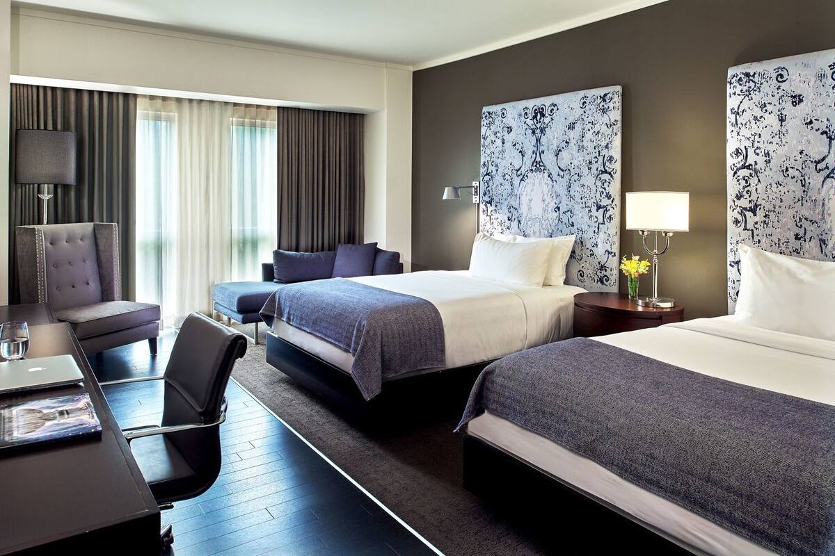 A hotel room with two beds, a desk, a chair, and a large window with curtains.