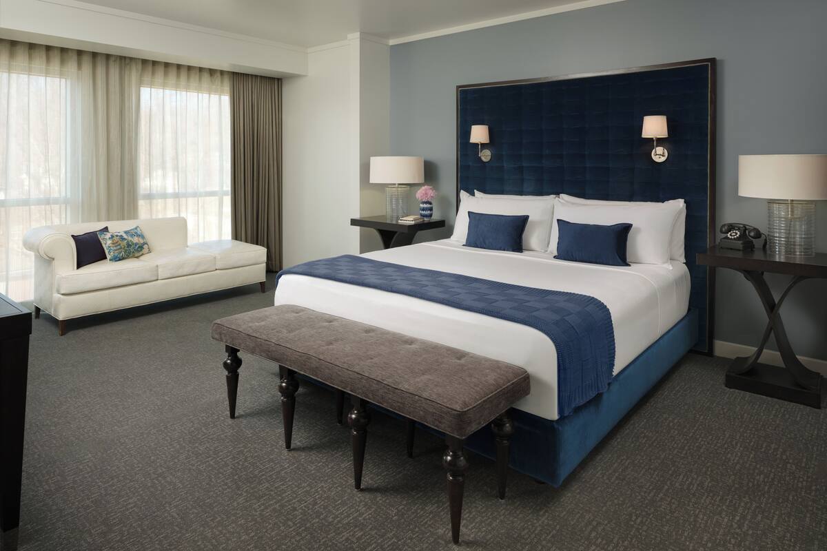 A hotel room with a large bed, a bedside table with a lamp, a small table with a lamp, a sofa, and a bench.