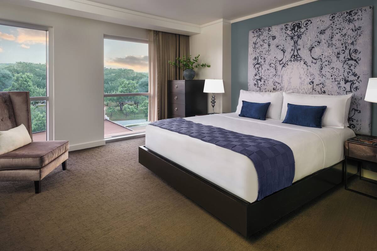 A hotel room with a large bed, a chair, a nightstand, a lamp, a dresser, and a view of the outdoors.