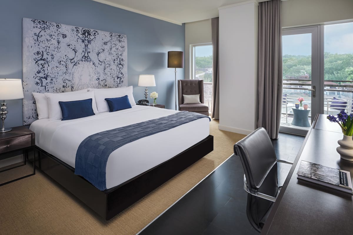 A modern hotel room with a large bed, a desk with a chair, a view of the city, and a large window.