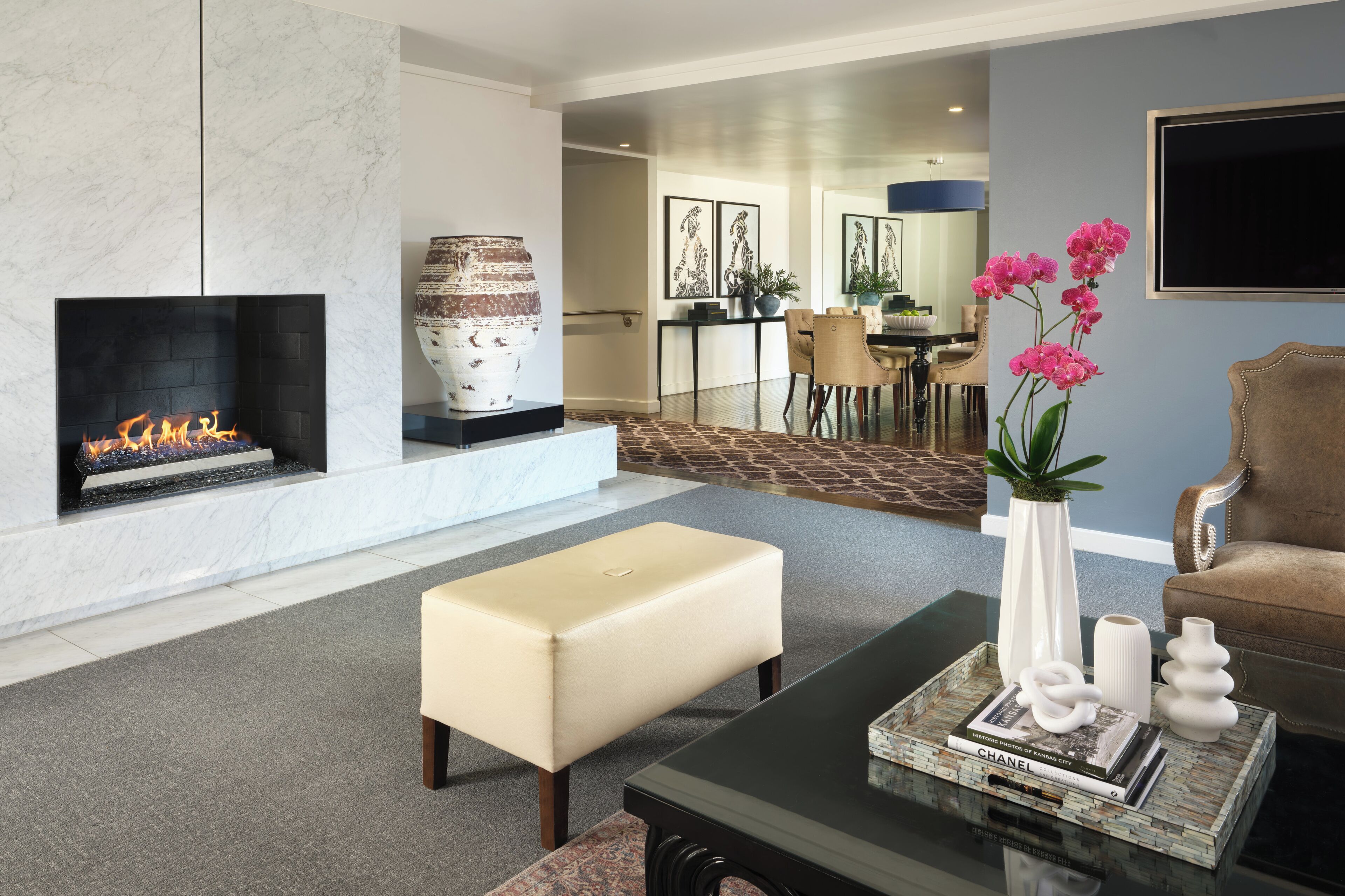 Una sala de estar con chimenea, una mesa de centro con libros y objetos decorativos, un reposapiés color beige, y un comedor al fondo.
