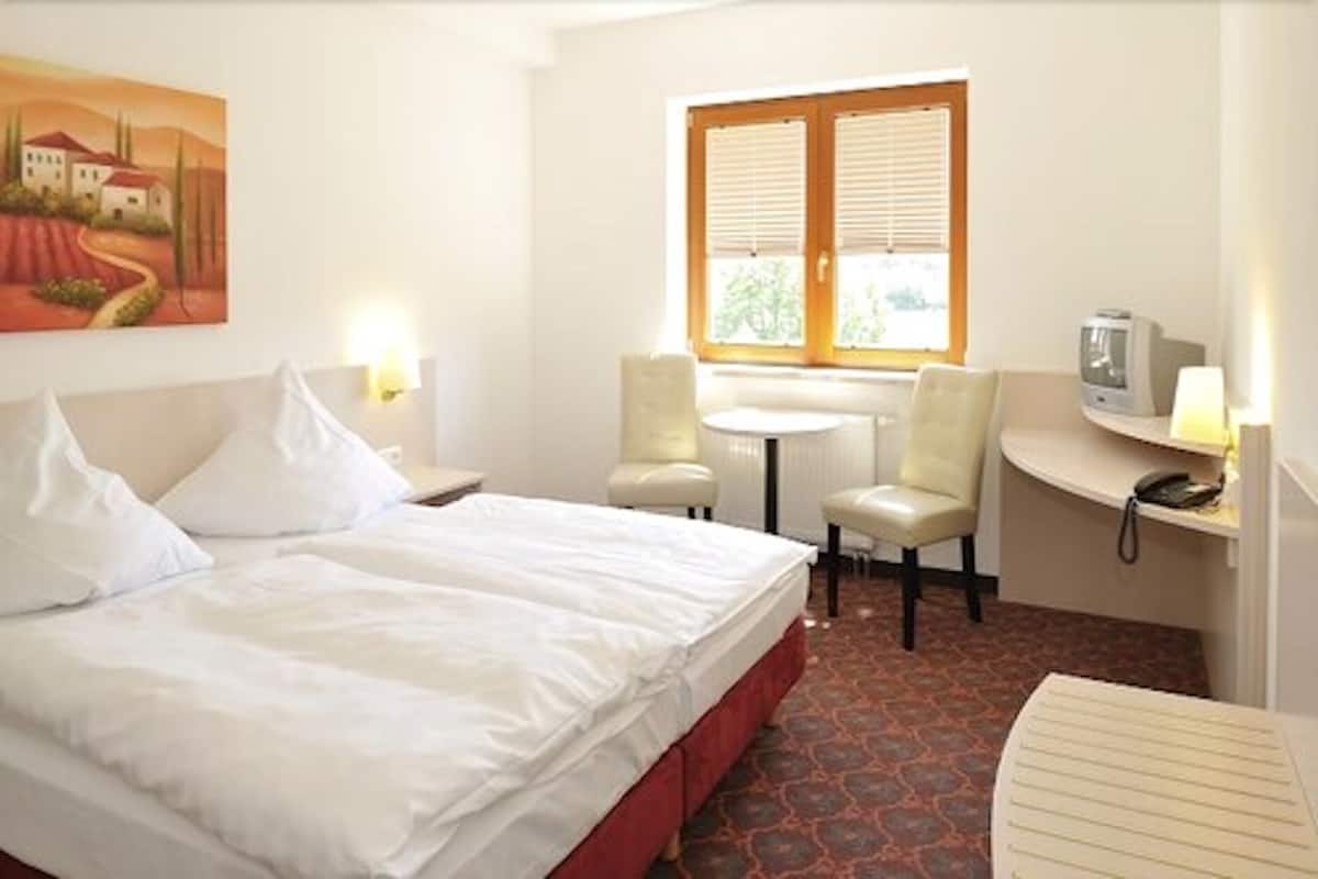 A hotel room with a bed, two chairs, a small table, a window with blinds, and a painting on the wall.