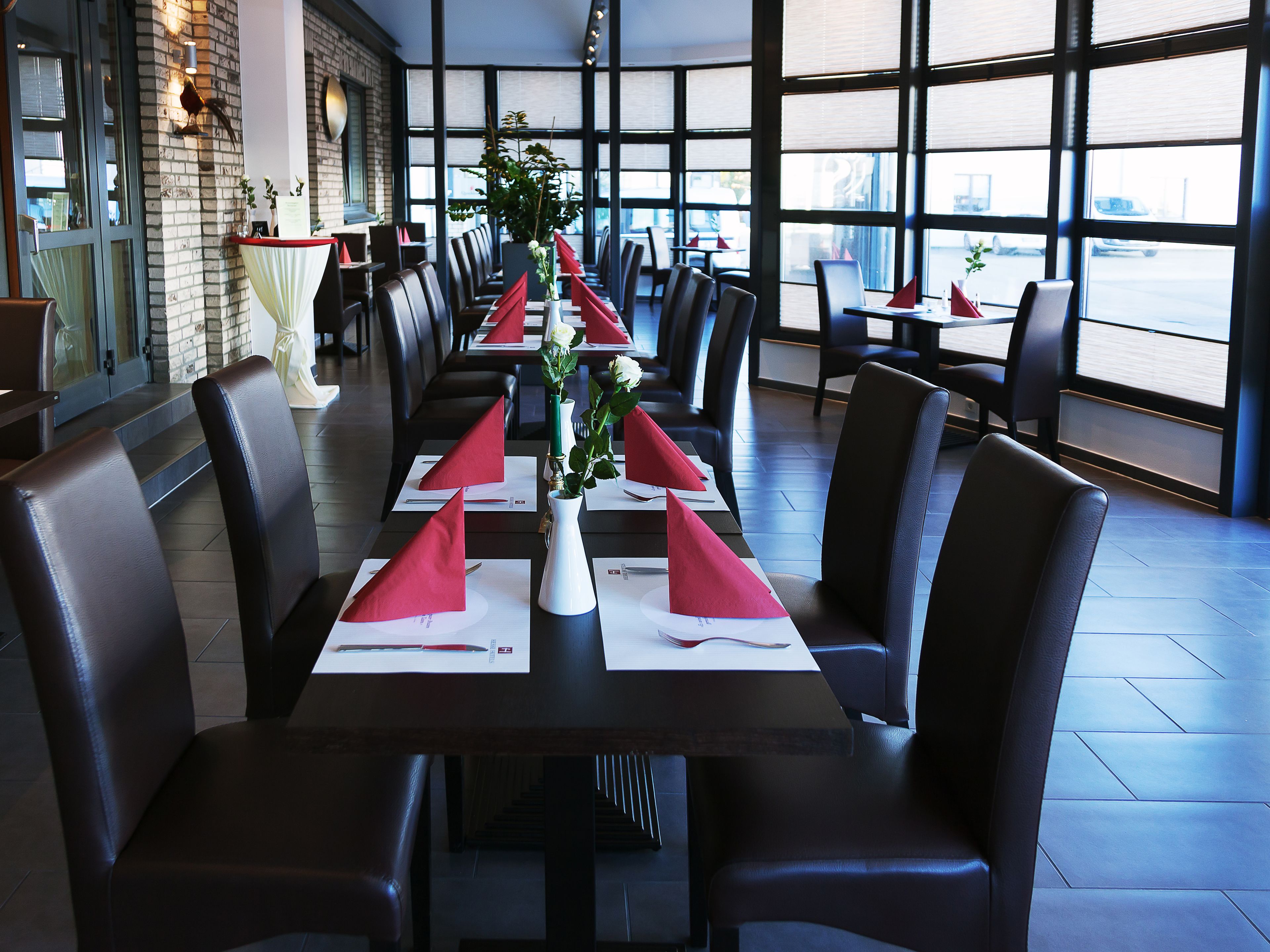 En restaurant med et langt bord dækket til måltid, med røde servietter, hvide tallerkener og en vase med blomster.