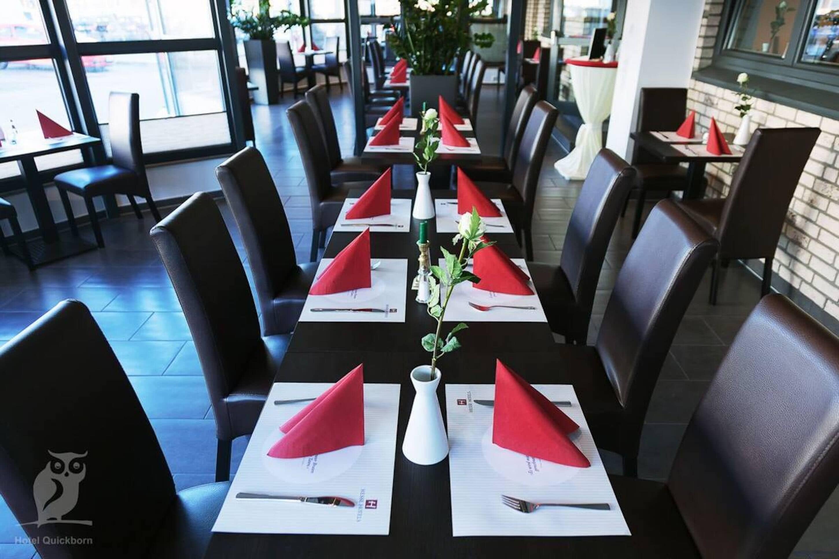 A restaurant with long tables set with red napkins, white placemats, and a vase with a plant.