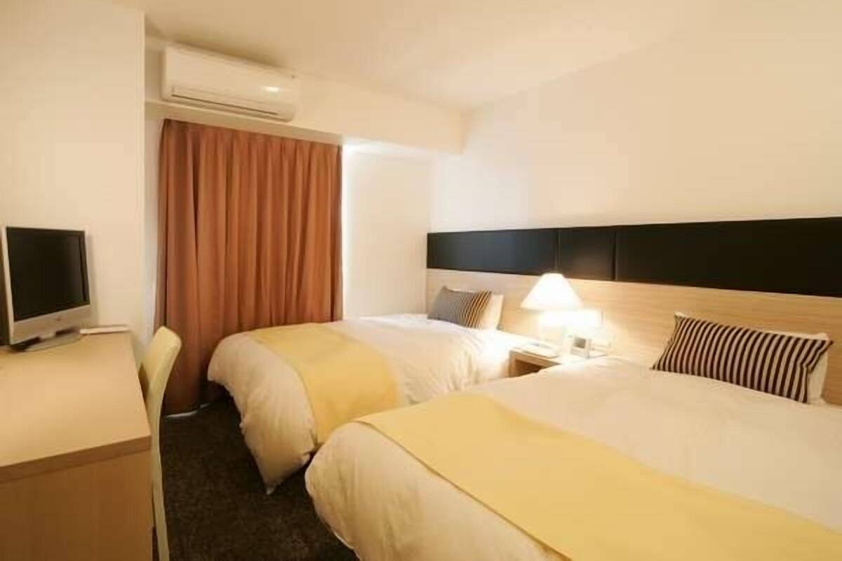 A hotel room with two beds, a desk with a computer, and an air conditioning unit.