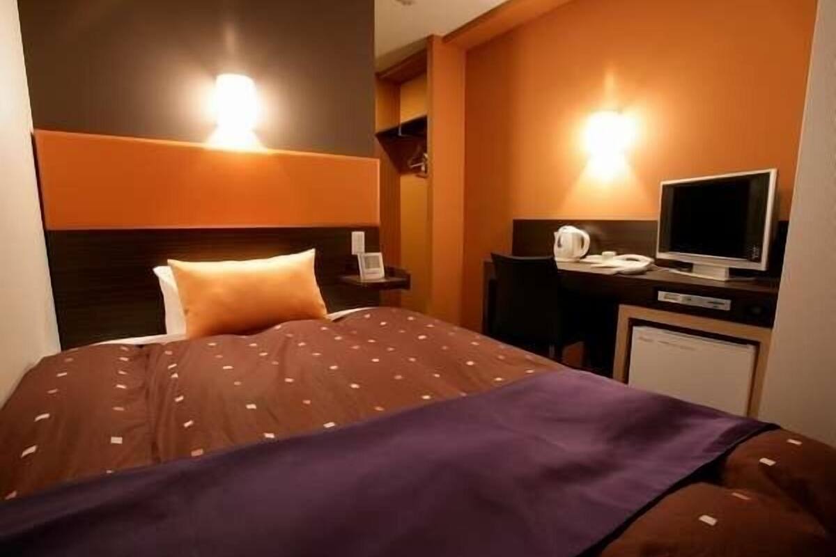 A hotel room with a bed, a desk with a computer and a kettle, and a television.