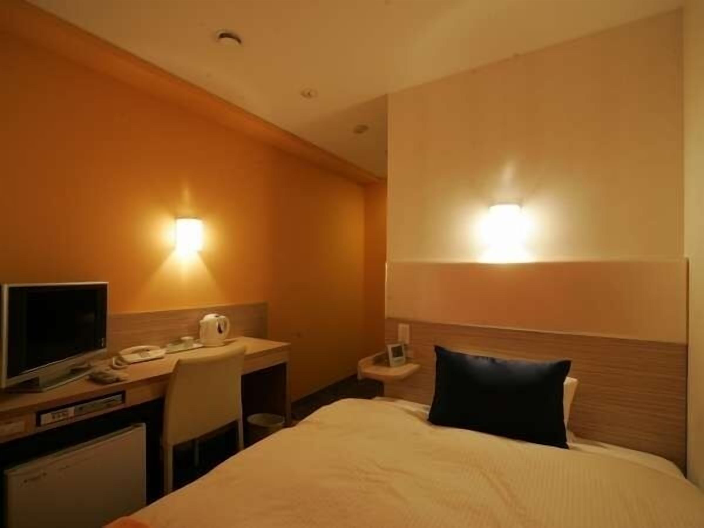 A hotel room with a bed, a desk with a computer, a chair, a nightstand, and a wall-mounted light fixture.