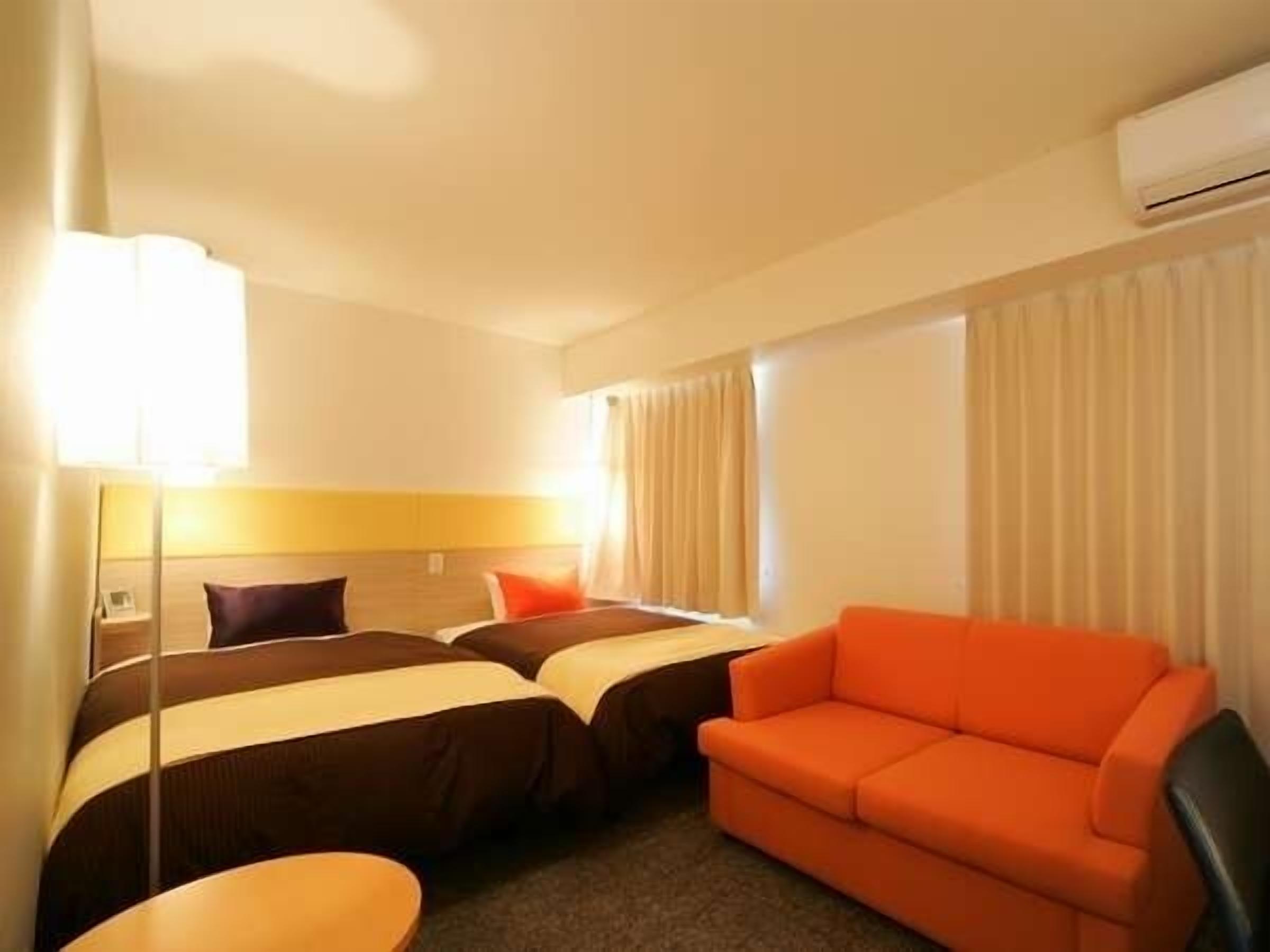 A hotel room with two beds, a sofa, a small table, and an air conditioning unit.