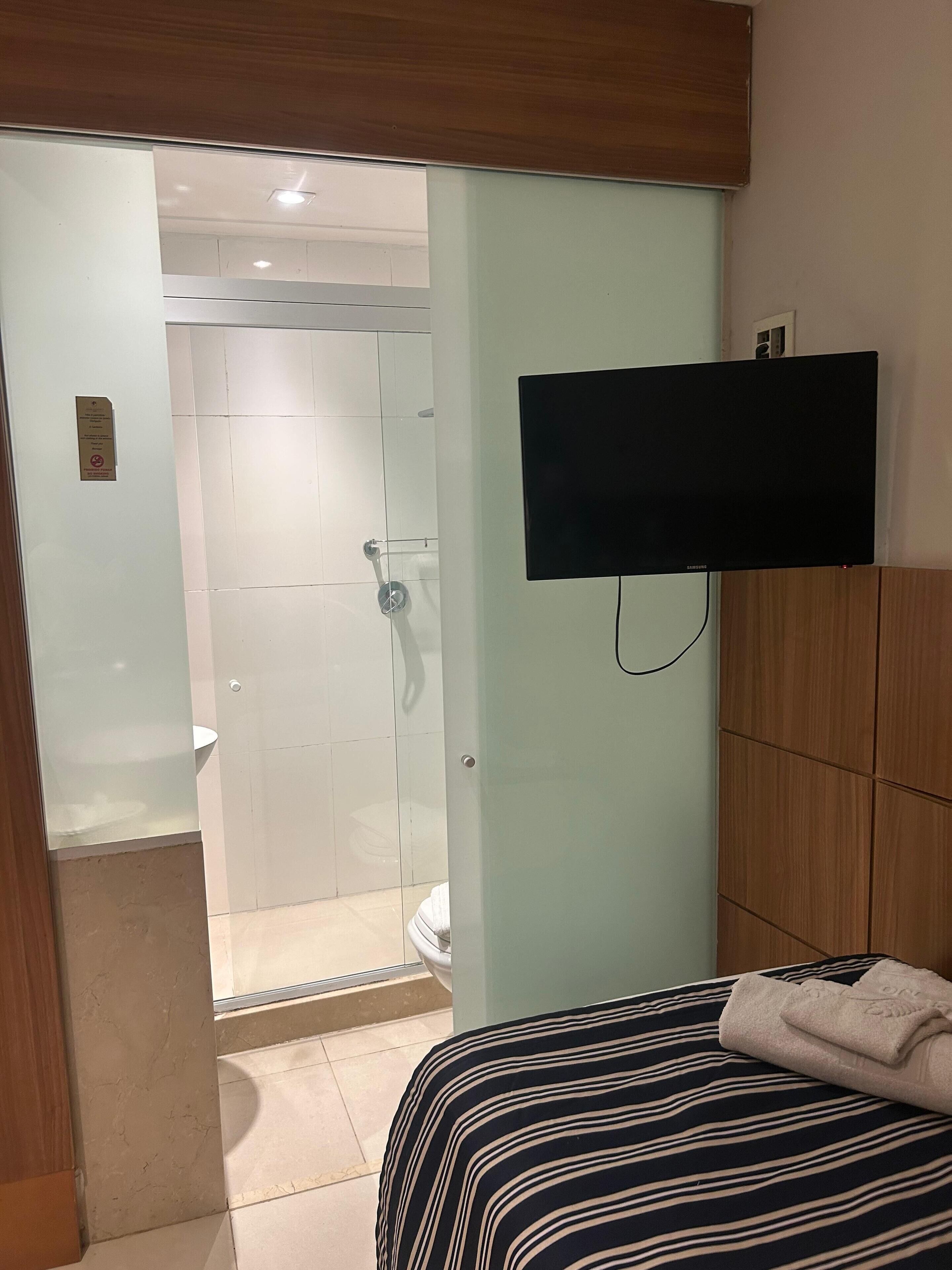 Habitación de hotel con cama, televisor montado en la pared, zona de ducha con paredes de cristal y cabecero de madera.