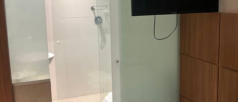 Habitación de hotel con cama, televisor montado en la pared, zona de ducha con paredes de cristal y cabecero de madera.