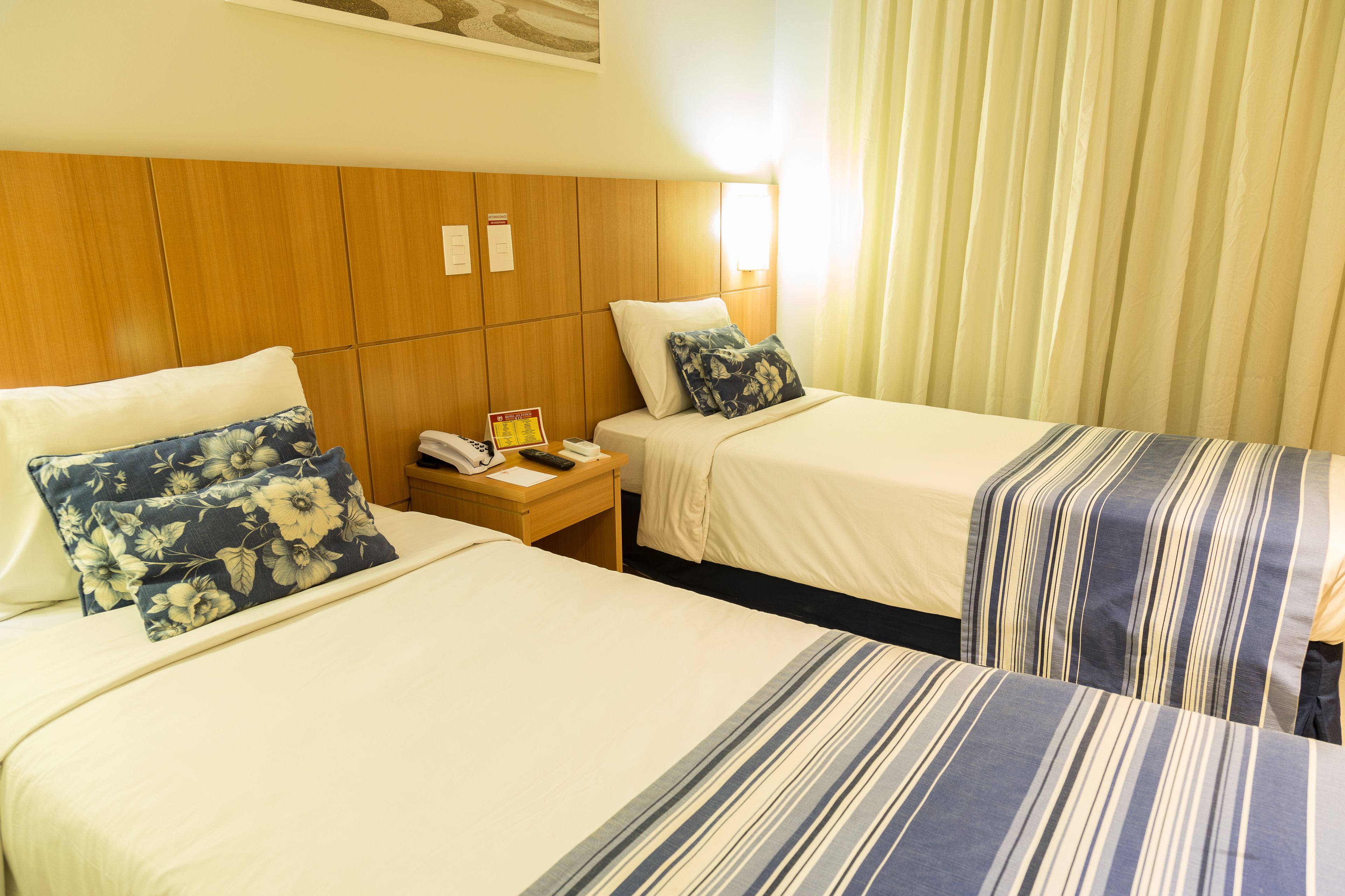 A hotel room with two beds, wooden headboard, bedside tables, and a curtain.