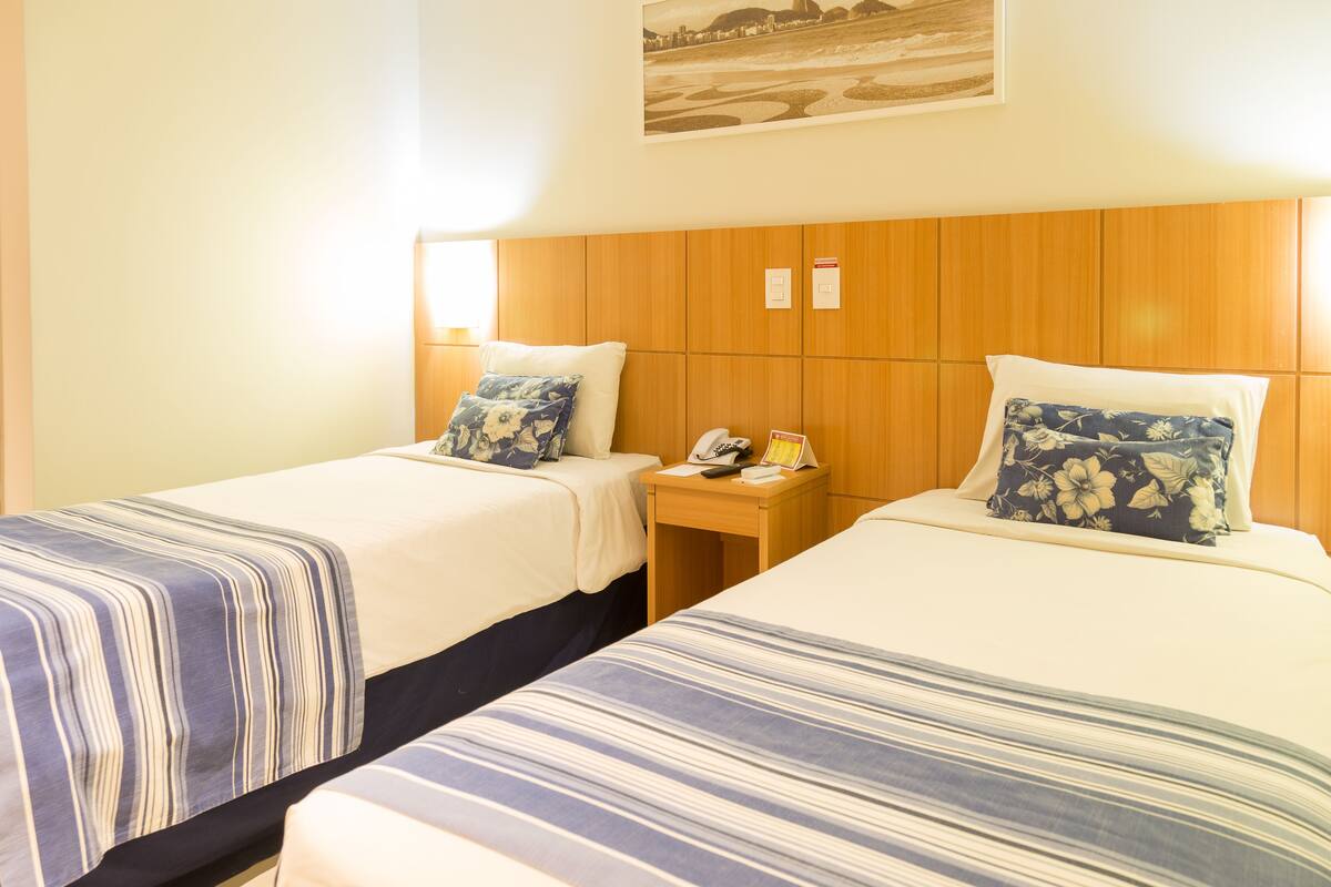 A hotel room with two beds, each with a striped bedspread, a wooden headboard, and a nightstand with a phone and a card.