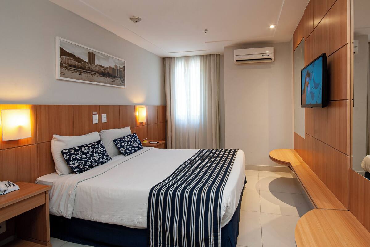 A hotel room with a large bed, a bedside table with a phone, a wall-mounted TV, and a painting of a cityscape.