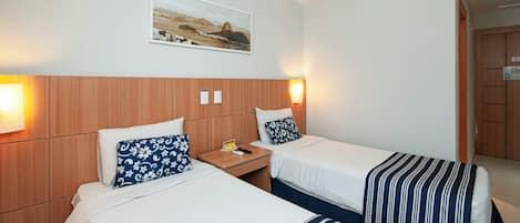A hotel room with two beds, a wooden headboard, a wall-mounted air conditioner, a framed picture, and a bedside table with a lamp.