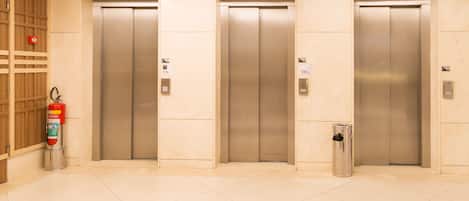 Three elevator doors in a lobby with a fire extinguisher and a trash can nearby.